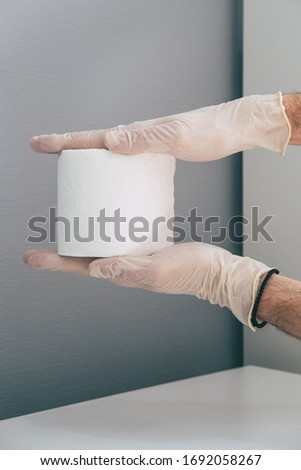 A medical worker in rubber latex gloves on his hands holds toilet paper roll . Concept of coronavirus panic. Grey and white colors. 