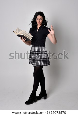 Portrait of a goth girl with dark hair wearing black and plaid skirt with boots. Full length standing pose, holding a book  on a studio background.