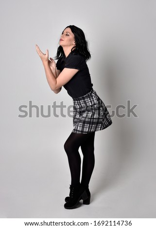 Portrait of a goth girl with dark hair wearing black and plaid skirt with boots. Full length standing pose on a studio background.