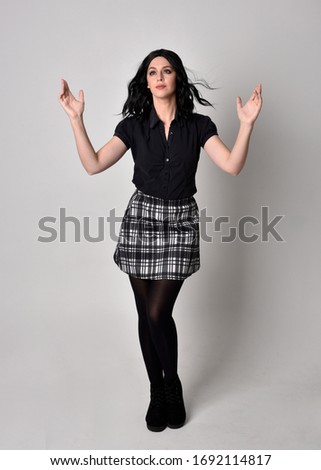 Portrait of a goth girl with dark hair wearing black and plaid skirt with boots. Full length standing pose on a studio background.