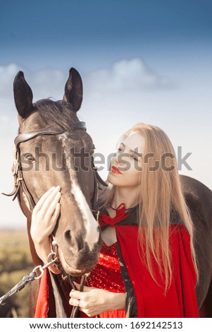 Girl blonde with long hair with a black stallion. In a red historical cloak