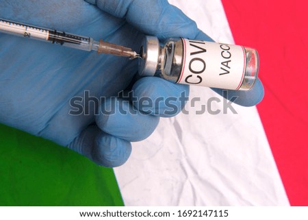 A doctor in blue rubber gloves holds an vial of the vaccine from COVID19 and a syringe against the background of the Italian flag. Fighting the epidemic.