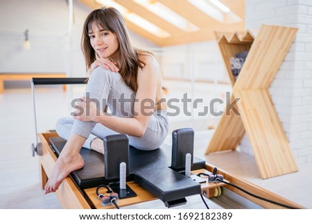 Portrait of female owner of small pilates studio wearing sportswear posing on sports equipment, reformer bed with light interior on background.