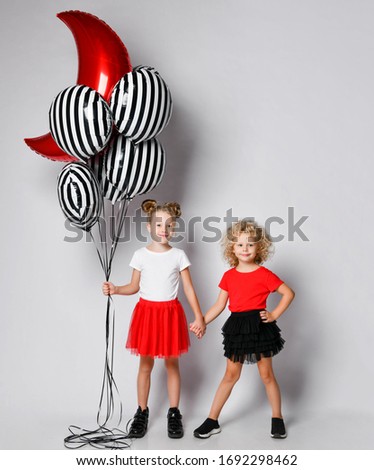 Two cute smiling blonde kids girls sisters in red and black skirts and white and red t-shirts stand together holding hands and big stylish air balloons