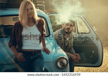 Young couple on a car trip. She leaned against the car's hood, he sits on driver's seat and smokes, selective focus on a man