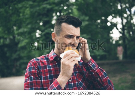 Handsome young man eating sandwich autdoor. He is holding a phone