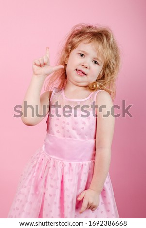 Little funny girl in a pink dress standing on a pink background showing thumb up