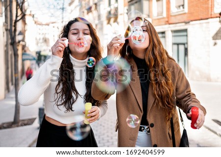 Stock photo of two young caucasian women blowing bubbles at the street. They are having fun together. They are wearing smart casual clothes.
