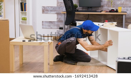 Professional worker in overalls consults furniture assembly instructions from laptop. Worker wearing a cap. Royalty-Free Stock Photo #1692413428