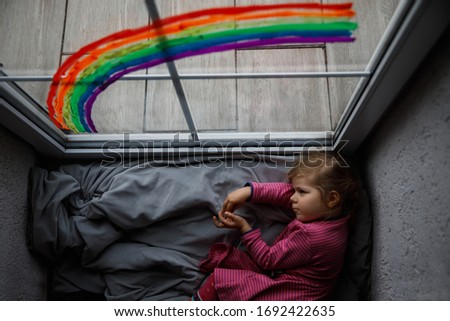 Adoralbe little toddler girl with rainbow painted with colorful window color during pandemic coronavirus quarantine. Child painting rainbows around the world with the words Let's all be well.