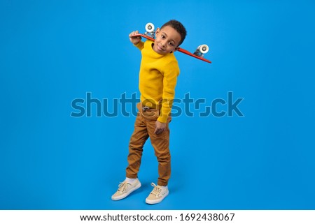 Cute African-American teenager in casual clothes yellow sweater and brown pants, walking and holding red skateboard and smiling against blue studio background. Concept of activity and happy childhood