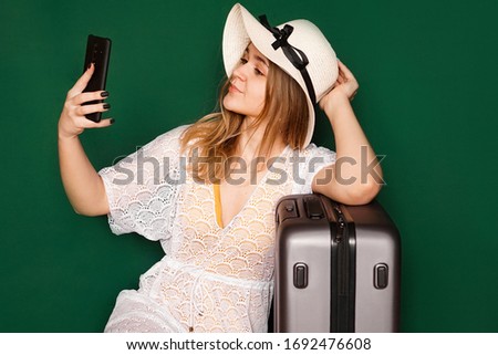 Elegant european woman taking selfie before vacation. Indoor shot of a lovely girl in a white dress sitting near her suitcase, green background