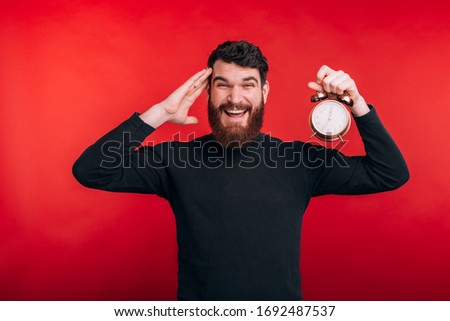 amazed bearded man standing over red background and holding alarm clockd