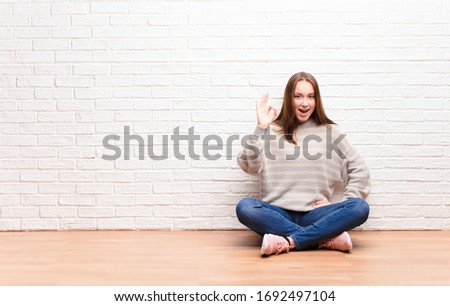 young blonde pretty girl feeling successful and satisfied, smiling with mouth wide open, making okay sign with hand sitting on the floor