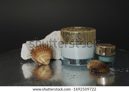 concept of skin care with marine ingredients jar of face cream and a small jar of cream under the eyes with drops of water and seashells on a dark background