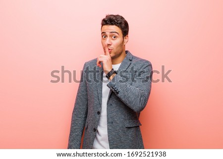 young arabian man asking for silence and quiet, gesturing with finger in front of mouth, saying shh or keeping a secret against pink wall