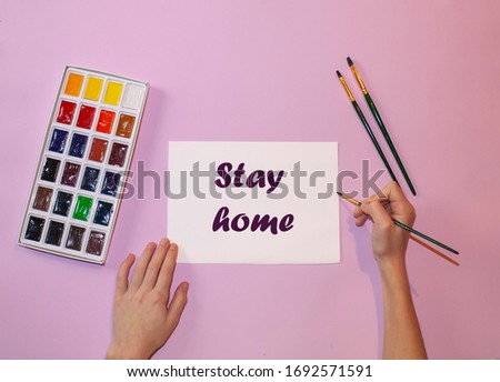 
a white sheet of paper with the words "stay home" on a pink background, next to a set of paints and brushes for painting