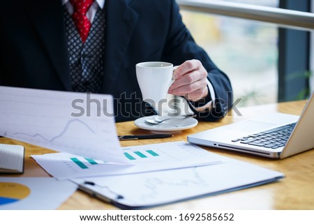 Adult male businessman working on a new project and looking at stock growth charts. Sits at a large window at the table. Looks at the laptop screen and drinks coffee.