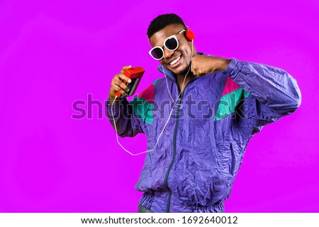 African American young man, in a jacket in the style of the 90s, with a retro cassette player, hears music, the mood of dancing and fun, yellow and purple colors Royalty-Free Stock Photo #1692640012