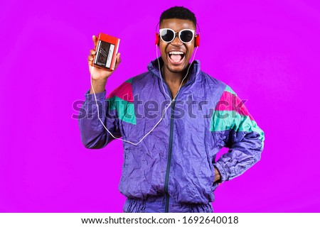 African American young man, in a jacket in the style of the 90s, with a retro cassette player, hears music, the mood of dancing and fun, yellow and purple colors Royalty-Free Stock Photo #1692640018