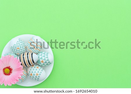 Decorated Easter eggs lie on the white plate with pink flower on green background. Happy Easter holiday and spring concept. Greeting, invitation card. Flat lay style with copy space.