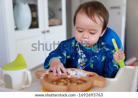 Cute baby boy learning how to use spoon eating yogurt by himself baby led weaning(BLW) self feeding on high chair at home. Mixed race Asian-German kid about 9-10 months old.Fine motor development. Royalty-Free Stock Photo #1692693763