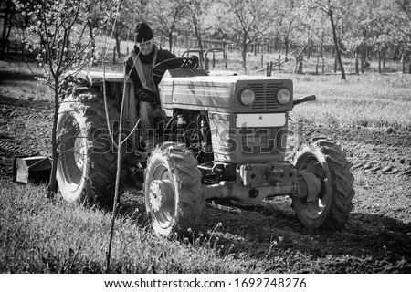 Old man works tilling the agriculture fields with his old rusty tractor. Old agricultural machinery tilling the soil. Farming and cultivation in Romania. Black and white picture.