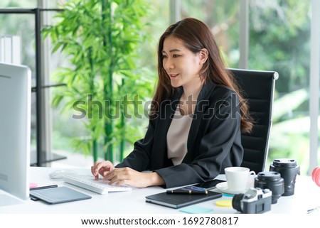 Asian business woman sitting in office looking at computer monitor with smile while camera and lens on her desk. Photography business concept.