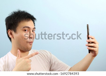 Thai mid age man with white shirt gesturing index finger pointing to mobile phone and expression happy "wow" looking to camera isolated in blue background. correct tick check mark symbol gesturing