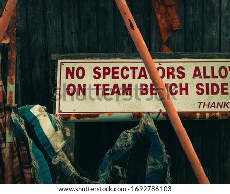 A close up photo of an small old barn at an empty local baseball field. On the side there is a rustic no spectators sign, and some ripped old fabric rapped on a rusted out metal bar.