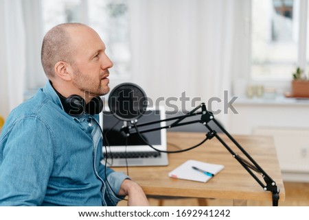 Man wearing earphones seated at a desk indoors alongside podcast equipment with a professional studio microphone and open laptop as he prepares to make a recording