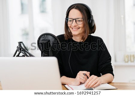 Smiling young woman in glasses on video conference call, using professional microphone and earphones, making notes in notebook. Remote worker during online meeting