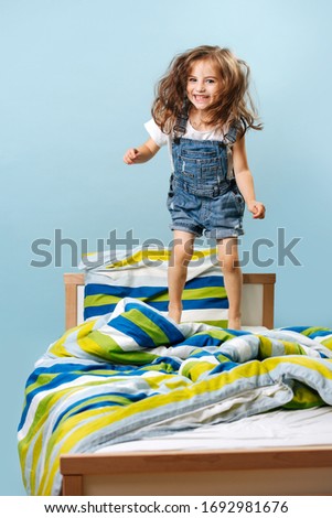 Naughty funny girl - toddler does not want to sleep, she jumps on the bed in a denim jumpsuit on a blue background