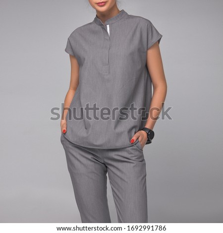 close-up of young stylish nurse in gray medical costume which is standing with hands in her pockets on gray wall background. medical concept. free space on left side