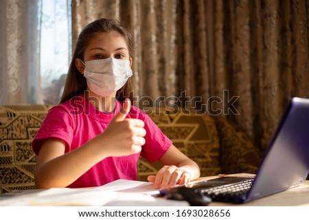 Young schoolgirl in medical mask showing thumbs up, happy to have classes at home, having distance learning and don`t go to school. Concept of life during coronavirus quarantine