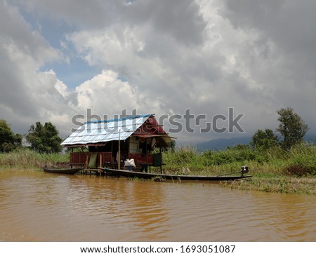 Landscape of Inle lake hut with boat parked outside. Maddy water. Tropical vegetation. Thick broken storm clouds. Trees. Myanmar in the summer.