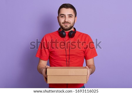 Front view of young delivery man with carton box in hands, posing isolated over lilac studio background, having headphones around neck, wearing red casual t shirt, looks at camera. Delivery concept.