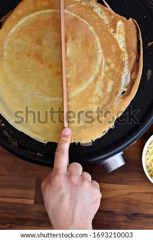 Picture taken from above - a person halves the dough on a crêpe iron. A man prepares delicious, homemade, sweet crepe by cutting the dough in half on the iron in front of a wooden background