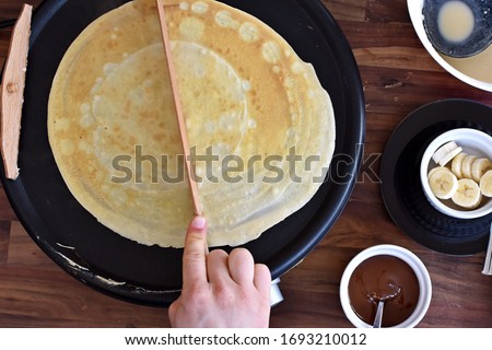 Picture taken from above - a person halves the dough on a crêpe iron. A man prepares delicious, homemade, sweet crepe by cutting the dough in half on the iron in front of a wooden background