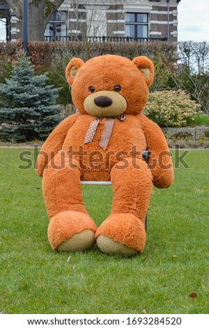 Stuffed huge bear sitting in the garden on a chair on green grass. A stuffed animal bear used for children to play with. Enormous stuffed animal, that has a brown color.