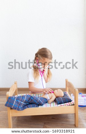 Little girl treats a doll, a child plays at the doctor in the room against a white wall, indoor