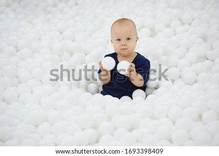  Cute child sits in  dry pool of white balls and plays looks at camera.