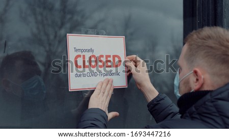Caucasian male wearing medical mask puts a Temporary closed due COVID-19 pandemic sign on a window. Coronavirus pandemic, small business shutdown Royalty-Free Stock Photo #1693447216