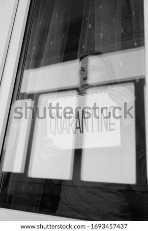 Woman looking through the window and holding a handwritten sign saying "Quarantine". Black and white portrait.