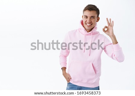Portrait of happy, satisfied male customer, guy with blond hair look pleased and smiling camera, show okay sign, say no problem, recommend good quality service, wear pink hoodie