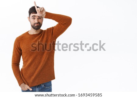 Portrait of funny handsome bearded man look gloomy, showing loser sign on forehead, feel let down and upset over failure, lacking confidence, standing white background uneasy