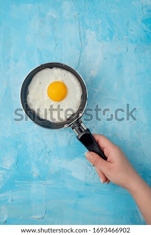 top view of woman hand holding frying pan with fried egg on blue background with copy space