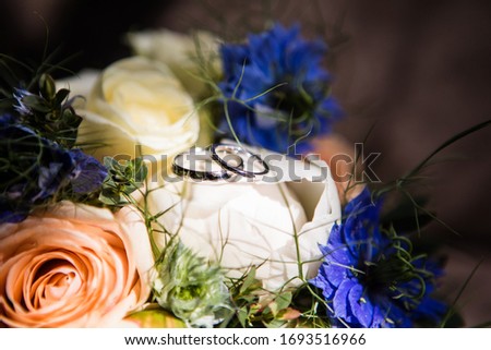 Macro picture of two white gold plain rings placed on the wedding bouquet with white peonies, cream and peach roses and bright blue knapweeds