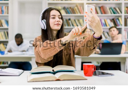 Attractive happy young girl student, wearing casual clothes and hipster earphones, sitting at the desk with books, holding mobile phone for selfie photo, while studying at the college library