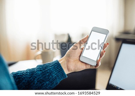 Closeup of a mobile cell phone of a young woman staying home and leadling business from distance on a freelance basis. Laptop, cup and flower in the room on the background
